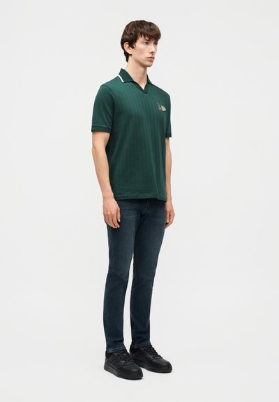 Νέος άνδρας που στέκεται, φορώντας ένα σκουροπράσινο πόλο μπλούζα με κοντά μανίκια και ένα μπαλώμα στο στήθος, σκούρα τζιν και μαύρα αθλητικά παπούτσια, μπροστά σε απλό φόντο.