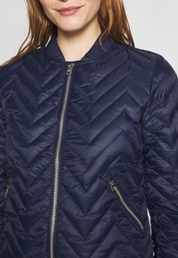 Marinblå kviltad bomberjacka med chevronmönster, ribbade krage och silverfärgade dragkedjor framtill och på fickorna. Lätt, vadderad textur.