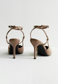Sandales à talons hauts en cuir verni beige avec une bride de cheville, présentant un bout pointu et un talon aiguille slim.