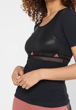 Kobieta z kręconymi włosami w czarnym sportowym topie Adidas Stella McCartney i różowych leginsach, z rękami złożonymi na talii.