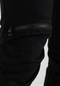 Svarta tygbyxor med en dragkedjeficka längs yttre lår. Dragkedjan är silverfärgad med en platt dragflik, vilket ger en funktionell detalj.