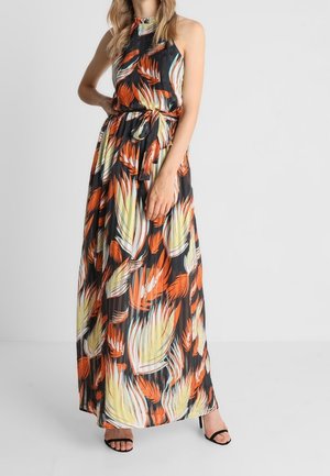 Žena v bezrukávých maxi šatech s vysokým límcem a vzorem tropických listů v oranžové, žluté a černé barvě, doplněná černými otevřenými podpatky.