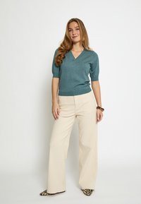 Maglione verde acqua a maniche corte con scollo a V abbinato a pantaloni color crema a gamba larga. La modella indossa scarpe nere e bianche con motivi e un braccialetto di legno.