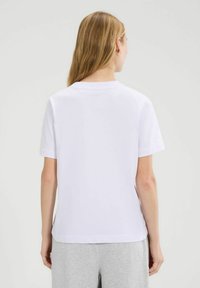 Vrouw met lang blond haar, gekleed in een eenvoudige witte short-sleeve t-shirt en lichtgrijze broek, met de rug naar de camera toe tegen een egale achtergrond.