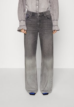 Persoon die vervaagde zwarte wijd uitlopende jeans en een grijs overhemd met knopen en gerimpelde manchetten draagt, staande tegen een effen achtergrond.