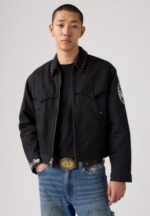 Jacket corta negra con tachuelas doradas en contraste, detalles bordados y cierre de cremallera. Combinada con pantalones de mezclilla azules y un cinturón decorativo.