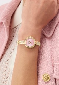 Gold bracelet watch with pink face and crystals worn on wrist over lace blouse and pink textured jacket with gold floral button.