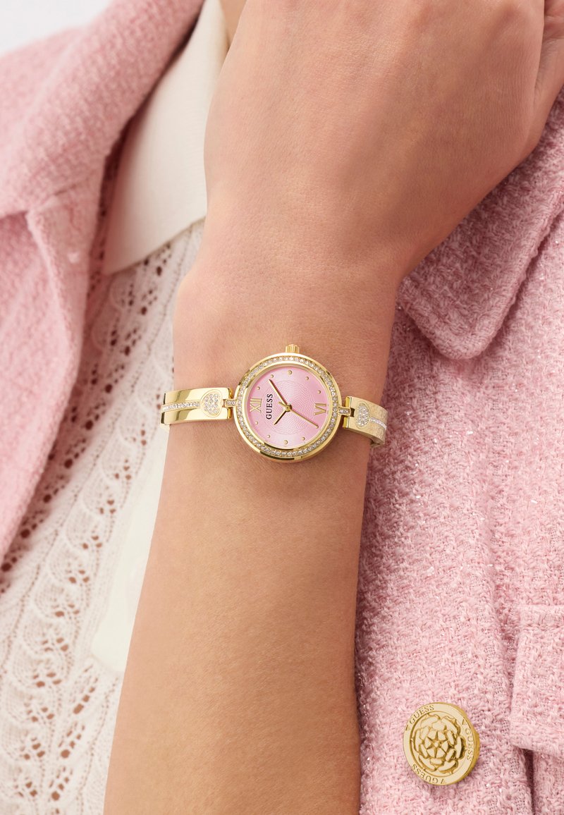 Gold bracelet watch with pink face and crystals worn on wrist over lace blouse and pink textured jacket with gold floral button.