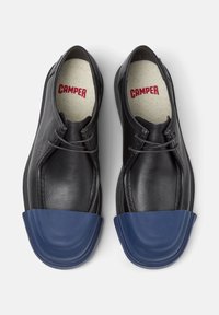 Zapatos negros con puntera de goma azul, con cordones y una plantilla texturizada con "CAMPER" impreso en rojo.