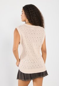 Pull sans manches en rose clair avec une texture côtelée et de petites ouvertures, porté sur une jupe en jean foncé avec un ourlet effiloché.