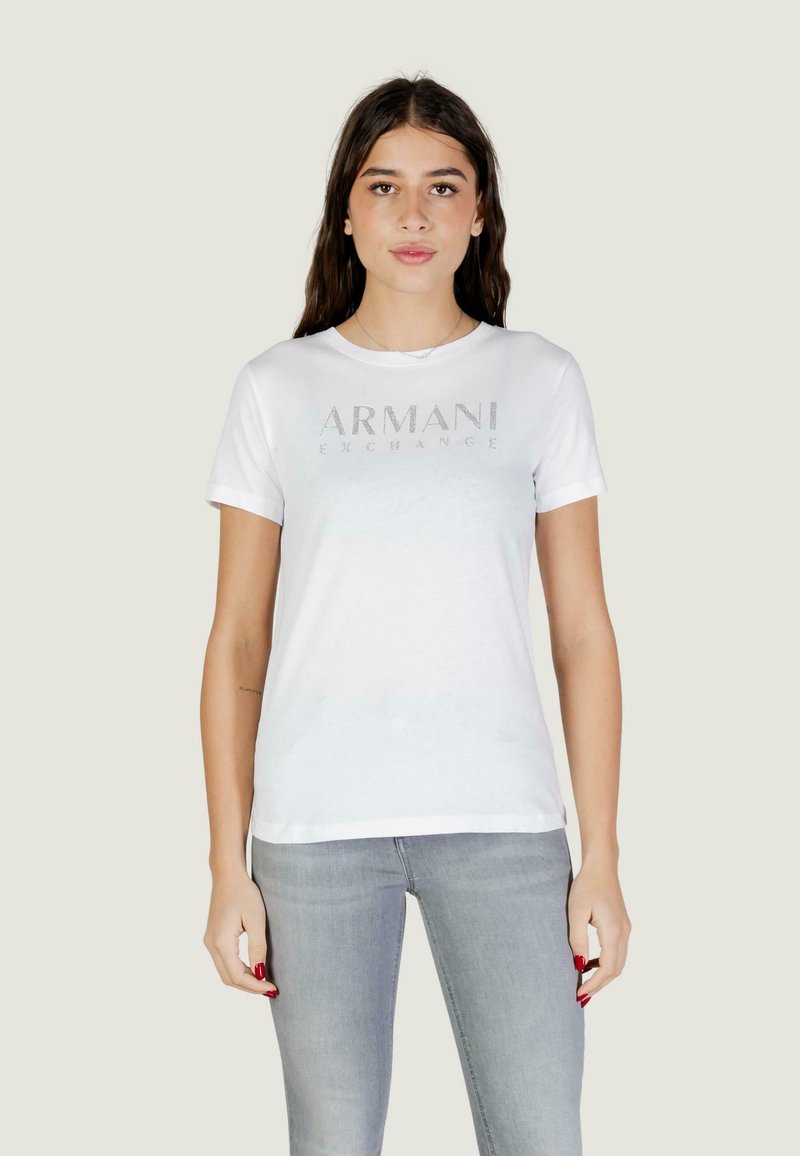 Jeune femme portant un t-shirt blanc Armani Exchange et un jean gris clair, debout devant un fond clair uni.