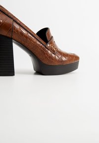 Mocassin plateforme en cuir marron à texture crocodile, avec un talon épais noir en bloc et un design sans lacets, présenté sur un fond blanc.