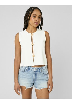 Femme aux cheveux tressés portant un haut blanc sans manches à boutons en forme de coquillage et un short en denim bleu clair, sur un fond gris uni.