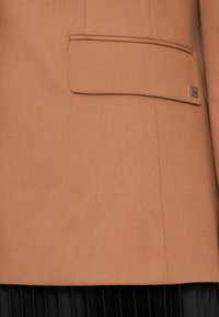 Blazer color cammello realizzato in tessuto texture, dotato di una tasca sul petto con un piccolo dettaglio di marca.