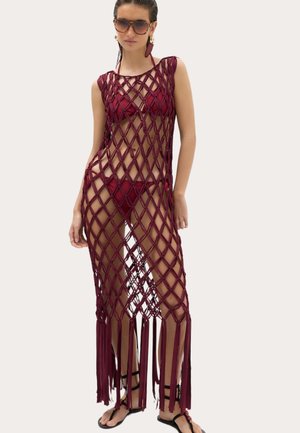 Kvinde iført vinrød bikini og lang vinrød netagtig kaftan med kvaster, solbriller og sorte sandaler mod lys baggrund.