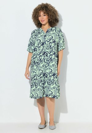 Frau mit lockigem Haar, die ein lockeres hellgrünes Kleid mit marineblauem Blumenmuster und graue flache Schuhe trägt, steht an einer weißen Wand.