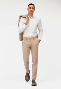 Camicia bianca a bottoni, pantaloni color beige chiaro, scarpe marroni. Il modello tiene una giacca color beige sulla spalla. Tessuto liscio, vestibilità sartoriale, palette neutra.