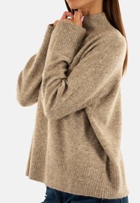 Suéter de punto beige con cuello alto, puños acanalados y un corte holgado. La textura parece suave, con un ligero patrón jaspeado.