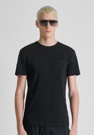 Hombre rubio con gafas de sol negras, camiseta negra lisa de manga corta y pantalones negros, sobre un fondo gris liso.