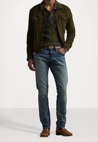 Chaqueta de ante verde oscuro sobre un polo verde oscuro, combinada con vaqueros azul claro y zapatos de cuero marrón. Unas gafas de sol reposan sobre la chaqueta.