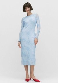 Robe à manches longues, bleu clair, avec broderies florales, silhouette ajustée et manches transparentes. Associée à des escarpins rouges à bout pointu.