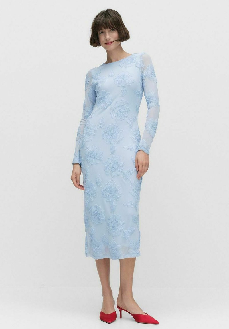 Robe à manches longues, bleu clair, avec broderies florales, silhouette ajustée et manches transparentes. Associée à des escarpins rouges à bout pointu.