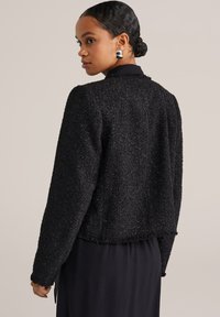 Naisel, kellel on juuksed kokku pandud bun'i, kannab hõbedasi kõrvarõngaid ja tekstureeritud musta pikakäelist jope musta kleidi üle, vaatab üle oma õla.