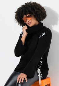 Schwarzer Strickpullover mit hohem Rollkragen, Glockenärmeln und weißen Schnürdetails an den Seiten. Getragen mit glänzenden schwarzen Leggings.