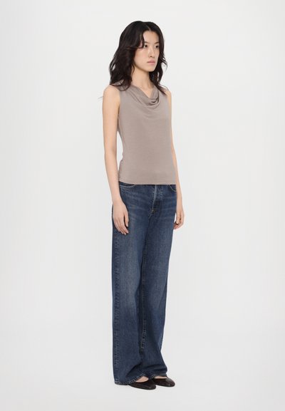 Ermeløs, grå topp med cowl-halslinje kombinert med vide, mørkeblå denimjeans. Modellen står foran en nøytral bakgrunn, iført svarte leiligheter.