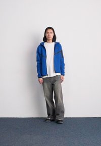 Casaco azul com fecho éclair e detalhes em preto, t-shirt branca, jeans largos desbotados, ténis pretos, em pé contra um fundo branco simples.