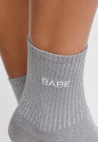 Calcetines grises de canalé con brillo plateado, con un logo blanco "BABE" en el puño. Vista cercana de la textura y forma de la tela.