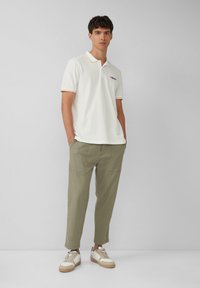 Weißes Polohemd mit orangefarbenen Akzenten, kombiniert mit olivgrünen tapered Hosen. Die Schuhe sind in Beige und Weiß gestaltet und verfügen über eine Gummisohle.