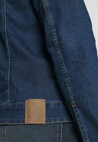 Dunkelblaue Jeansjacke mit sichtbaren Nähten. Mit einem braunen Leder-Label auf der Rückseite. Glatte Textur und traditionelles Design.