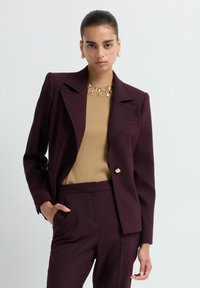 Femme portant un costume violet foncé avec un haut beige orné d'embellissements métalliques au niveau du col, se tenant devant un fond blanc uni.