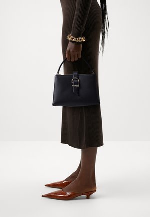 Femme en robe marron côtelée tenant un sac à main noir, portant un bracelet épais en or et des chaussures marron à talons bas pointues.