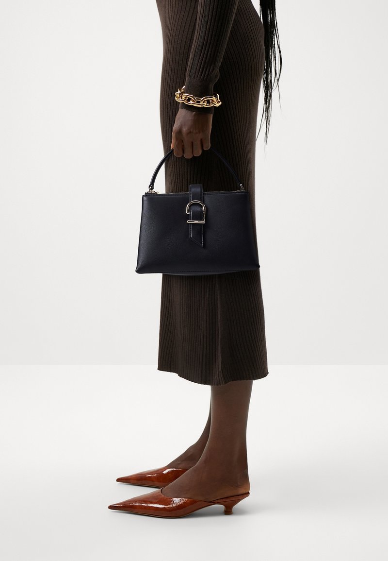 Femme en robe marron côtelée tenant un sac à main noir, portant un bracelet épais en or et des chaussures marron à talons bas pointues.