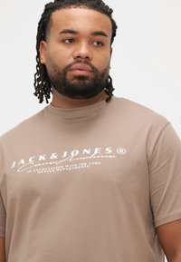 Ruskea puuvillainen T-paita, jossa valkoinen painettu "JACK & JONES" -logo rinnassa, pyöreä pääntie ja lyhyet hihat.