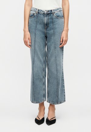 Femme portant un jean taille haute à jambes larges bleu avec des coutures verticales proéminentes, associé à des escarpins noirs à bout pointu et à bride arrière.