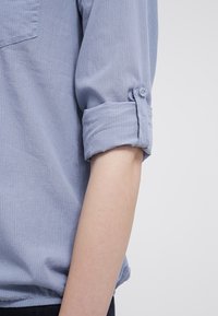 Chemise bleu clair à manches longues en tissu texturé. Manches retroussées maintenues par des boutons. Comprend une poche poitrine et une coupe décontractée.
