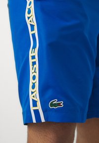 Μπλε σορτς φτιαγμένα από υφασμάτινο ύφασμα διαθέτουν λευκή πλευρική ρίγα με το "LACOSTE" σε κίτρινο χρώμα και ένα πράσινο κροκόδειλο στο στρίφωμα.