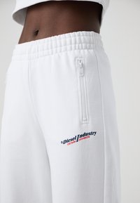Baltas sporta bikses ar gludu tekstūru, elastīgu jostasvietu, ar rāvējslēdzēju sānu kabatu un "Diesel Industry Denim Division" logo sarkanā un zilā krāsā.