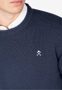 Suéter de punto azul marino con cuello redondo, que presenta un logotipo bordado en blanco de utensilios cruzados en el pecho. Detalle de tejido texturizado visible.