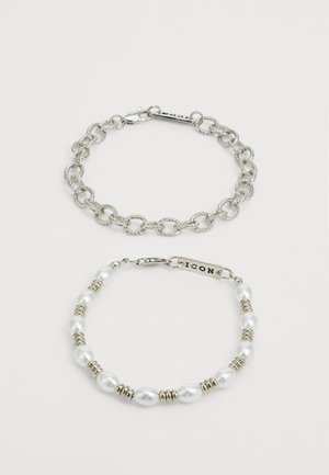 Strieborný náramok z reťazových článkov s prepojenými článkami nad náramkom s korálkami, ktorý obsahuje biele perly a kovové medzičlánky, zaistený zapínaním.