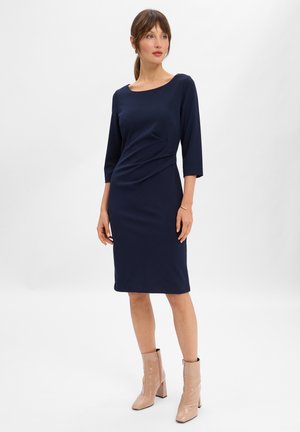 Marineblaues Kleid mit Dreiviertelärmeln, gerafftem Detail an der Taille, knielangem Saum, kombiniert mit beigen Lackstiefeln bis zum Knöchel.