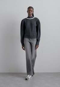Une femme avec des cheveux tressés porte un pull gris foncé à manches bouffantes, un jean gris, des baskets noir et blanc, et un collier de perles blanches.