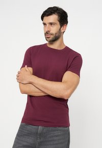 Mann mit dunklem Haar und Bart, trägt ein kastanienbraunes T-Shirt und graue Jeans, die Arme verschränkt, schaut leicht zur Seite vor einfarbigem Hintergrund.