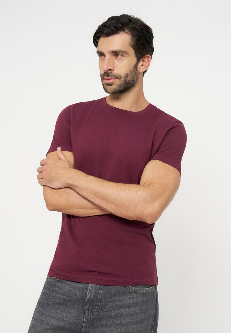 Man met donker haar en baard, draagt een bordeauxrood T-shirt en grijze spijkerbroek, armen gekruist, kijkt iets opzij tegen een effen achtergrond.