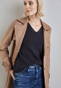 Hellbrauner, strukturierter Trenchcoat über einem dunkelblauen V-Ausschnitt-Pullover, kombiniert mit blauen Jeans. Glatte Stoffe und taillierte Passform.