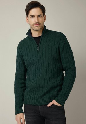 Zielony sweter w sploty z półzamkniętym kołnierzem, grubymi ściągaczami na rękawach i dole oraz teksturowanym wzorem, zestawiony z ciemnymi dżinsami.