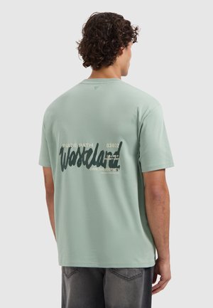 Man met krullend haar die een lichtgroen T-shirt draagt met de tekst "Wasteland" en reisgerelateerde tekst op de achterkant, staand tegen een effen achtergrond.